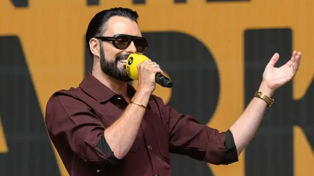 Rylan Clark speaks into a microphone on a stage with his other hand out to the side of his body. He wears a burgundy shirt, sunglasses, gold watch and bracelet.