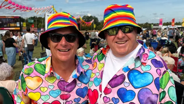 Two men wear matching multi-coloured bucket hats and multi-coloured shirts with love hearts on them in different colours. They both wear sunglasses and are smiling at the camera.