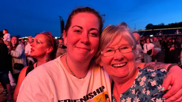 A woman with dark hair which is tied back who wears a white T-shirt, hugs an older woman with grey hair and glasses who is wearing a floral top.