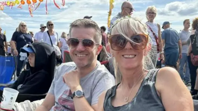A man and women sit next to each other on camping chairs. They both wear sunglasses. The man has short dark hair and wears a grey T-shirt. The woman has blonde hair that has been tied up and she wears a dark grey vest top.