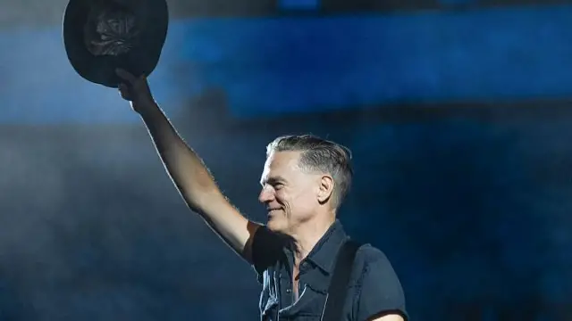 Bryan Adams raises his hat in the air on stage during a performance. He looks out on to the crowd and smiles.