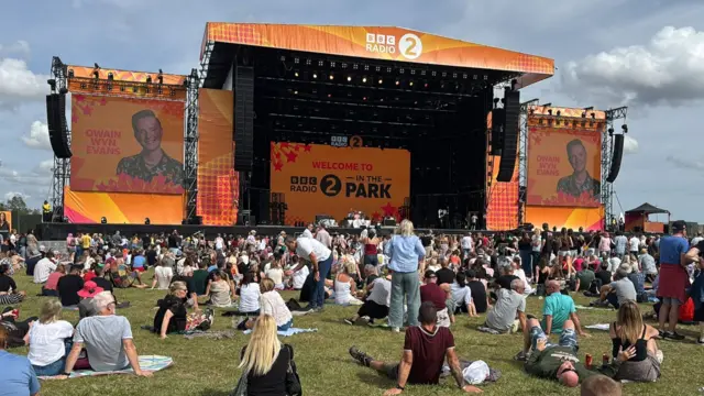 The large main stage at the festival. It is orange and covered in orange BBC Radio 2 branding. Lots of people are either standing in front of it or sitting on the grass.
