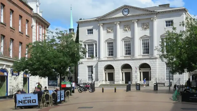 This image shows the top of Chelmsford High Street, which has a cafe and a bookmaker. The most prominent feature is Shire Hall, a large white building of three levels. It is grand in its appearance, with large windows and tall columns.