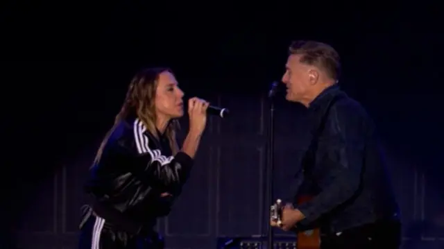 Mel C and Bryan Adams sing to each other on stage. Bryans plays a guitar as Mel C sings into a microphone in her hand.