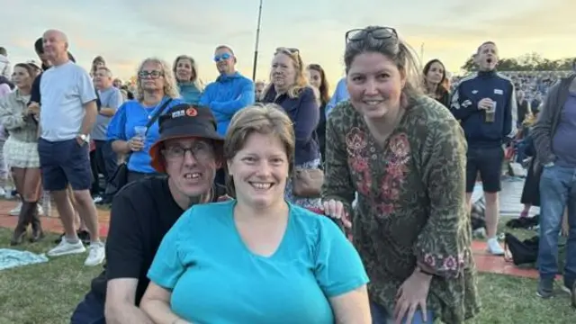 A man wearing a bucket hat and black T-shirt kneels next to a woman wearing a blue T-shirt in a wheelchair. Another woman stands to their right in a green floral long top and jeans.