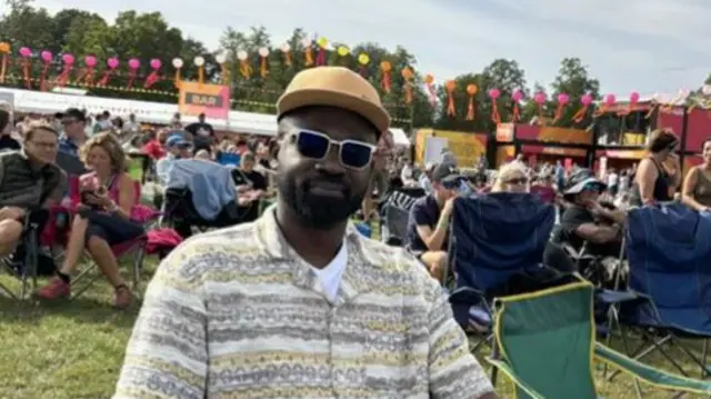 A man sits in a camping chair at a festival surrounded by other attendees. He wears a beige cap, sunglasses and a patterned shirt with a white T-shirt underneath.