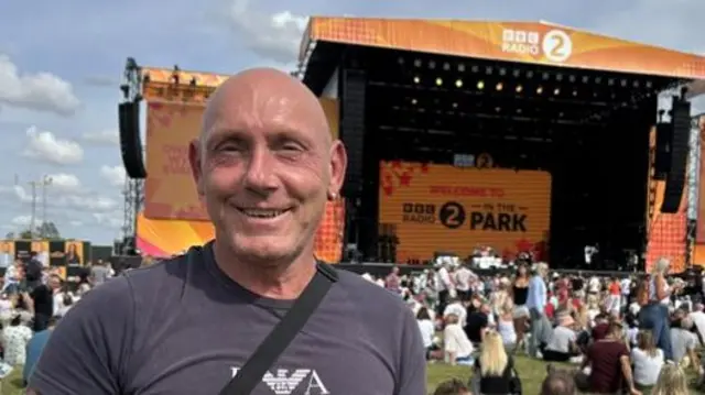 Mark Talbot is wearing a grey T-shirt and is smiling in front of the large orange main stage.