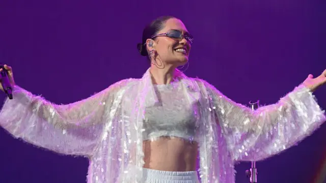 Jessie J raises both her arms and smiles during a performance.She wears a silver glittery cropped top and trousers as well as a jacket over the top.