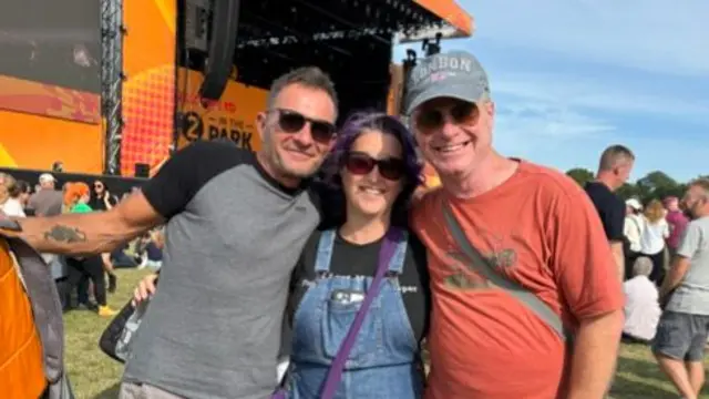 A woman with purple hair stands between her two male friends. They are all wearing sunglasses. The woman wears a denim dress with a black top underneath. One man wears a grey T-shirt and the other wears an orange one.
