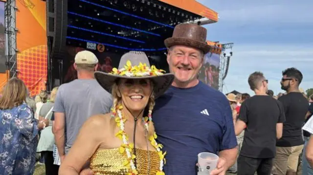 A woman wears a grey cowboy hat with yellow flowers on it and yellow flowers around her neck. She also wears a gold dress. A man stands next to her wears a brown top hat and blue Adidas sports top. He holds a plastic pint cup in one of his hands.