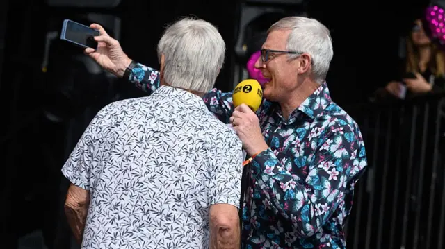 Tony Blackburn and Jeremy Vine take a selfie on a stage. They both wear floral shirts.