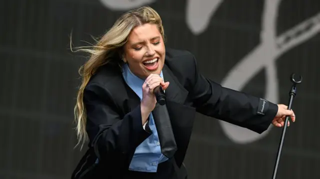 Ella Henderson during her performance at Radio 2 in the Park. She sings into a microphone in one hand while her other hand holds the microphone stand. She has long, curled blonde hair and she wears a black jacket, blue shirt and black trousers.