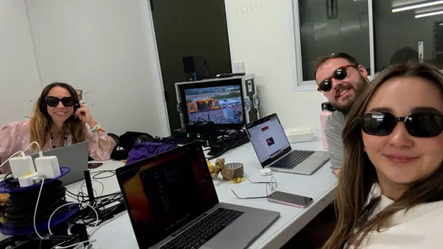 Two women and a man sit behind desks with laptops in front of them in a press tent at a festival. They are all wearing sunglasses, with one of the women wearing headphones.