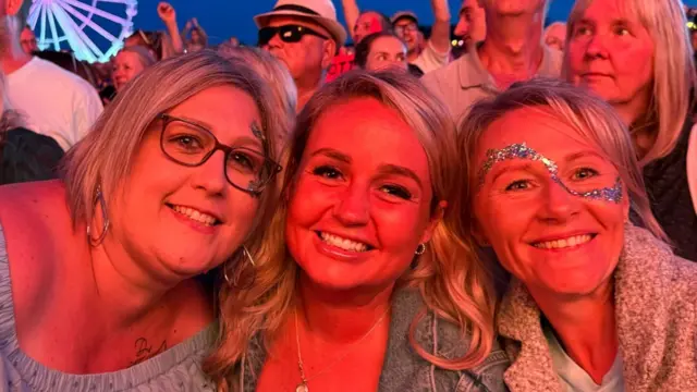 Three women all with blonde hair smile at the camera. Two women on the outside have face paint and one wears glasses. Crowds of people can be seen behind them.