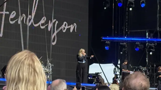 Ella Henderson stands on stage pointing out at the crowd. She has long blonde hair and wears a black jacket and trousers.