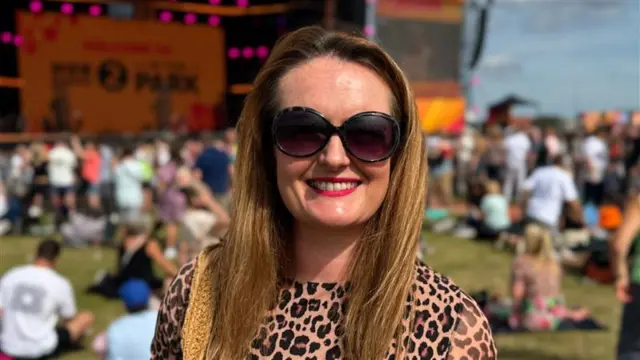 A woman with long light brown hair smiles at the camera in front of a large outdoor stage. She wears sunglasses and a leopard print top.