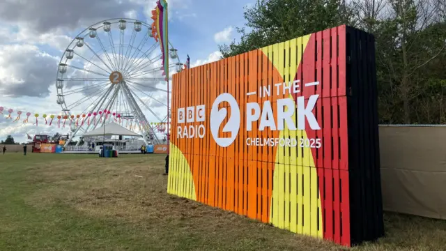 A large yellow, red and orange sign says "BBC Radio 2 in the Park, Chelmsford 2025". It is on grass in a field and there is a large ferris wheel in the background behind it.