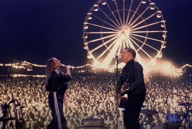 Mel C and Byran Adams sing to each other on stage. A large crowd can be seen behind them along with a ferris wheel.