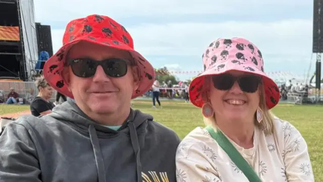 Philip and Jenny are wearing red and pink bucket hats respectively and sunglasses. Philip is wearing a grey hoodie and Jenny a cream long-sleeved top. They are sitting on the grass and smiling.