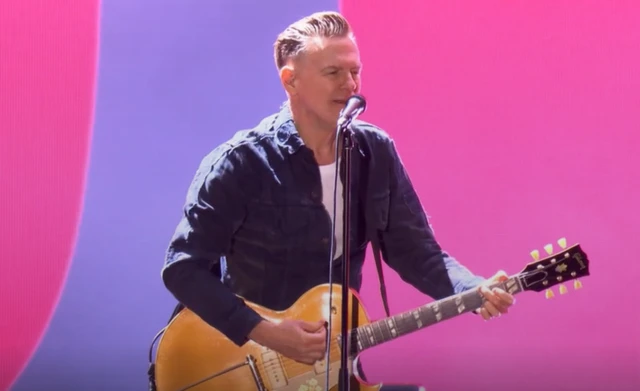 Bryan Adams performs on stage playing a guitar. He has short hair and he wears a blue jacket with a white T-shirt underneath.