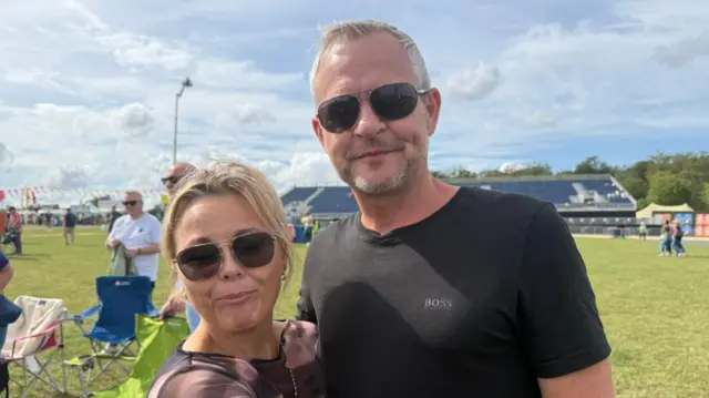 A woman with blonde hair that has been tied up and a man with short grey hair, wearing a black T-shirt smile at the camera. They both wear sunglasses.