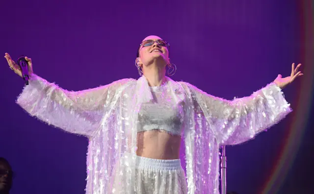 Jessie J looks up with her arms spread wide from her body. She wears a silver, glittery co-ord outfit with a cropped topped and trousers.