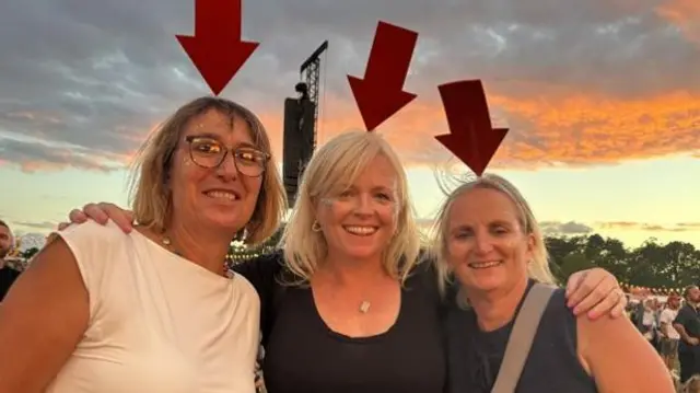 Three women smile at the camera. They wear matching hats that are a large red arrow pointing down at their heads.