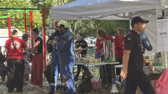 A crowd of people, including some in red t-shirts and some in blue emergency services uniforms, gather round a gazebo