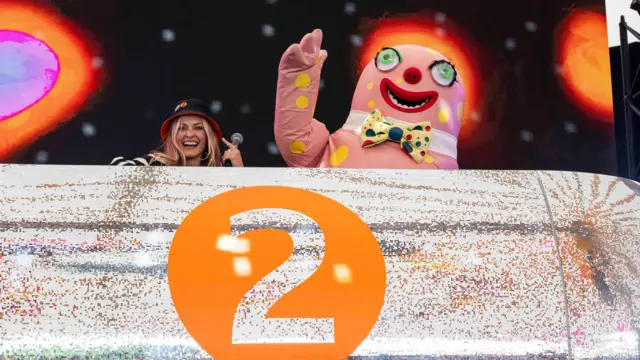 Mr Blobby in his pink outfit with yellow dots dances behind a glittery silver DJ desk. He is next to Fearne Cotton, who wears a black bucket hat and is smiling.