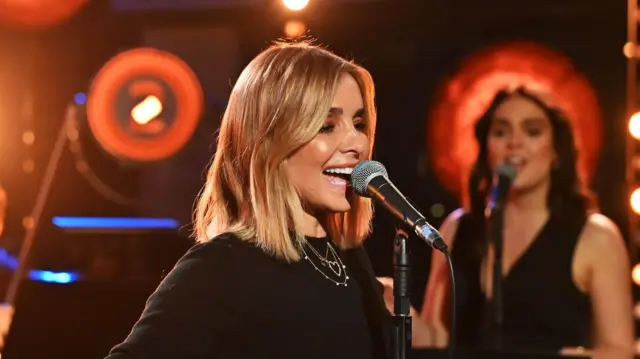 Louise performing a Radio 2 Piano Session in 2023. She has medium length blonde hair and is wearing a long sleeves black top, black pants and a necklace. Her arms are stretched out to either side of her as she sings. Behind her are backing singers and other musicians. The lighting is a soft orange colour.