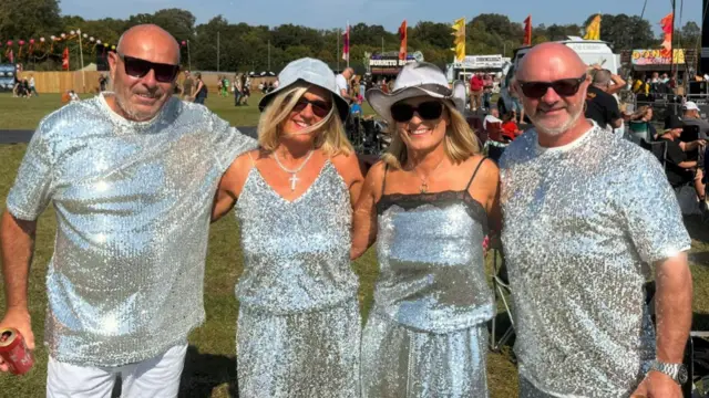 Two women wearing matching outfits that are silver and sparkling stand in between two men wearing similar tops. They all wear sunglasses and smile at the camera.