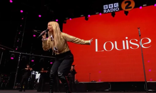 Louise performs on stage. A large screen behind her has her name written on it against a red backdrop. She has long blonde hair and wears a gold jacket with black shorts and knee high boots.