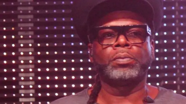 Jazzie B is standing in front of a wall of lights as he performs. He is wearing thick square framed glasses, a large black hat and a grey top with blue and orange sequins on. He has long dark hair that has been braided over one shoulder and grey and dark facial hair.