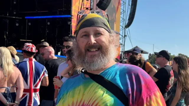 Jason Collins is wearing a colourful tie-dye T-shirt and a bandana. He is smiling and standing among the crowd near the main stage.