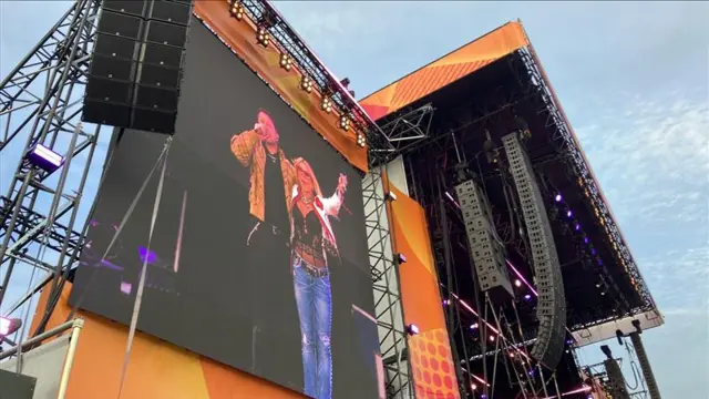 A large festival stage. Two performers can be seen on a large screen on the side of the stage.