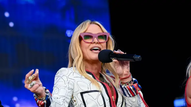 Anastacia sings into a microphone on a stage. She has long blonde hair and wears purple glasses along with a white jacket.