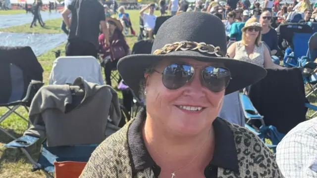 Bonnie Stobbs is wearing sunglasses and a black hat. She is smiling while sitting on a camping chair, in front of many others doing the same thing.