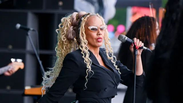 A woman with long blonde curly hair performs on stage. She wears glasses and a black cropped blazer. She holds a microphone in one hand.