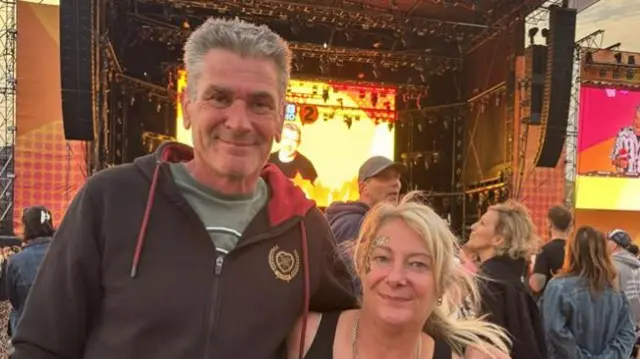 A man with grey hair, wearing a dark hoodie, has one arm around a woman with blonde hair that is tied up who wears a black vest. A large festival stage can be seen behind them along with other crowd members.