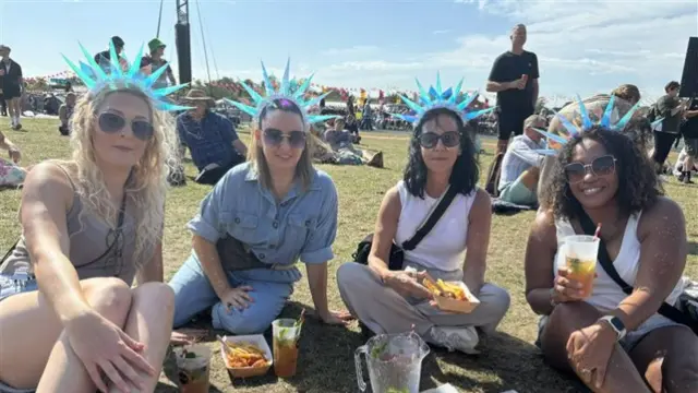 Four women sit together in a field. They wear matching hats that have blue spikes on them. They have food and drinks in front of them and they are all wearing sunglasses.