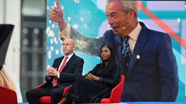 Healey and Badenoch sit in red chairs and look ahead while an image of Nigel Farage is projected on the screen behind them.