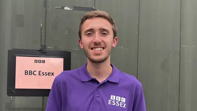 A young man wearing a purple polo top with the BBC Essex logo on the chest smiles at the camera. He has short brown hair and stands in front of wooden building with a poster on it that reads, BBC Essex.