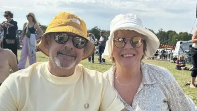 A man with a yellow T-shirt, bucket hat and sunglasses sits on the ground with a woman wearing a white bucket hat, sunglasses and silver sequin top.