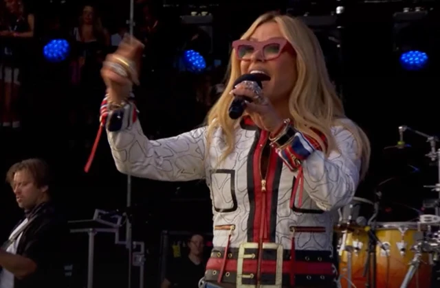 Anastacia sings into a microphone with her other hand raised in the area. She has long blonde hair and wears a white jacket with red and black details.