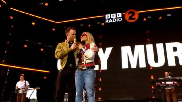 Olly Murs has his arm wrapped around singer Anastacia on a large stage. They are speaking into microphones and smiling.