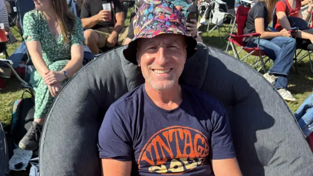 A man sits in a camping chair wearing a bucket hat and blue T-shirt. He smiles at the camera.