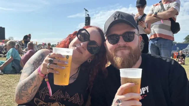 A woman with red hair and sunglasses wears a Def Leppard top and holds a pint of beer. She rests her head on a man who is also holding a pint of beer and is wearing sunglasses, a black T-shirt and black cap.