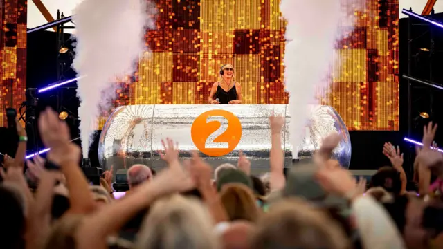Jo Whiley behind a silver DJ desk, with smoke cannons firing upwards either side of her. The people crowd in the foreground have their arms raised.
