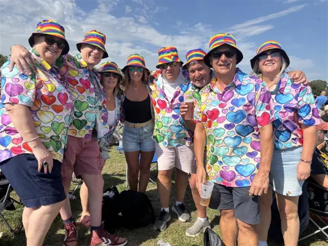 A group of eight people wear matching shirts with multicoloured shirts on and bucket hats. They smile at the camera and many of them are wearing sunglasses.