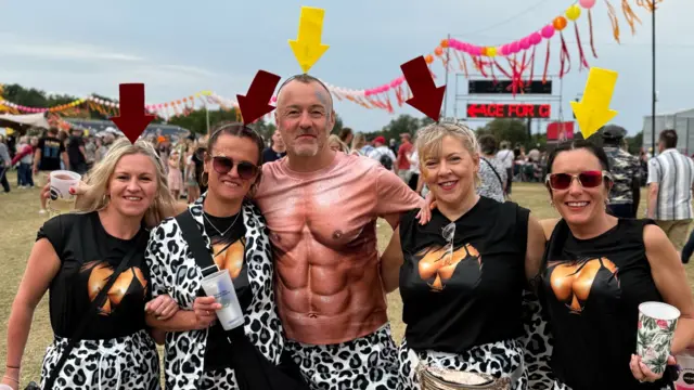 A man wearing a top with abs drawn on to it stands in between two women either side of him. The women wear matching tops and hats.